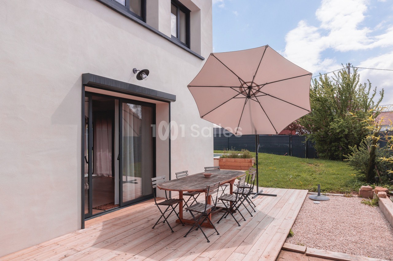 Terrasse en bois avec table, chaises et parasol beige, jouxtant une maison moderne et un jardin verdoyant.