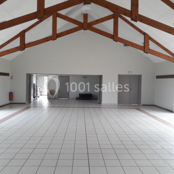 Salle vide avec carrelage clair, poutres apparentes au plafond et plusieurs portes grises sur les murs.