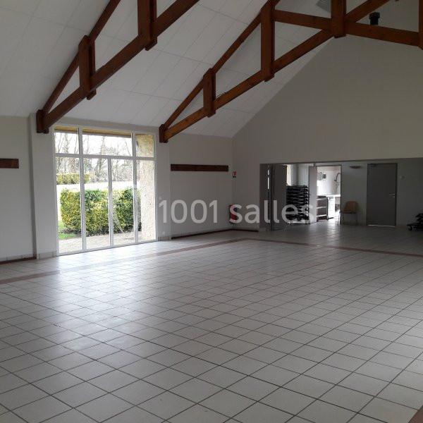 Salle vide avec carrelage clair, poutres apparentes, grandes baies vitrées et portes donnant sur un espace extérieur.