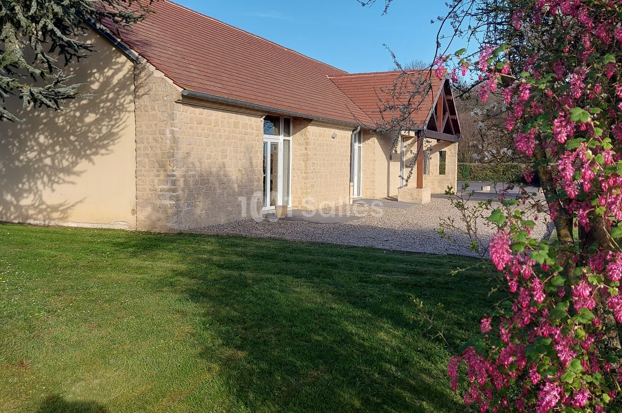 Bâtiment en pierre avec toit en tuiles, entouré de pelouse et d'arbustes fleuris sous un ciel dégagé.