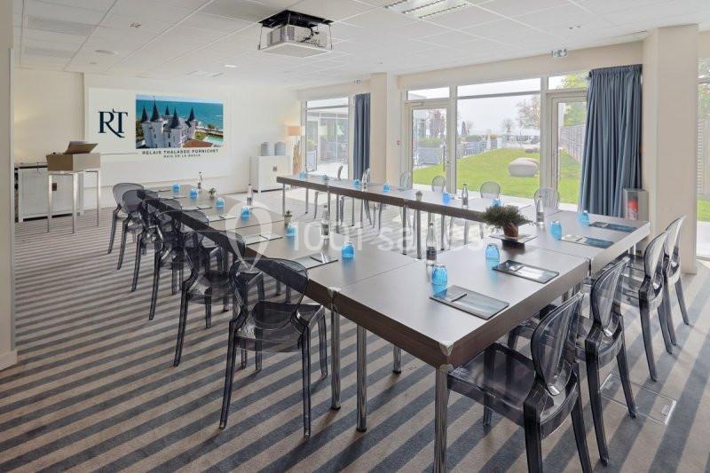 Salle de réunion lumineuse avec tables disposées en U, chaises transparentes et vue sur un jardin par de grandes baies…