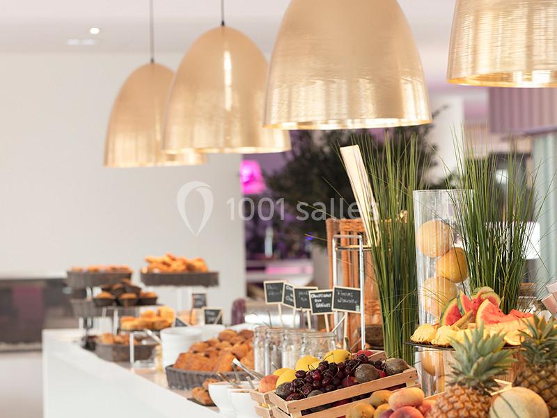 Buffet avec fruits frais, viennoiseries et pâtisseries, éclairé par des suspensions dorées dans un espace lumineux.