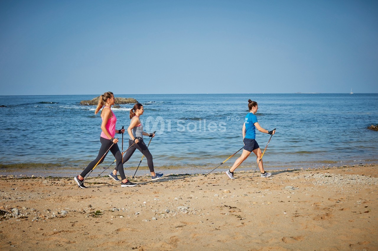 Trois personnes pratiquent la marche nordique sur une plage, avec la mer et le ciel bleu en arrière-plan.