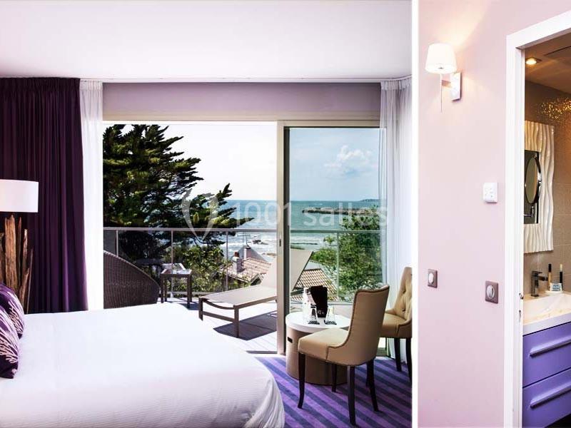 Chambre lumineuse avec vue sur la mer, balcon aménagé, fauteuils et salle de bain attenante visible à droite.