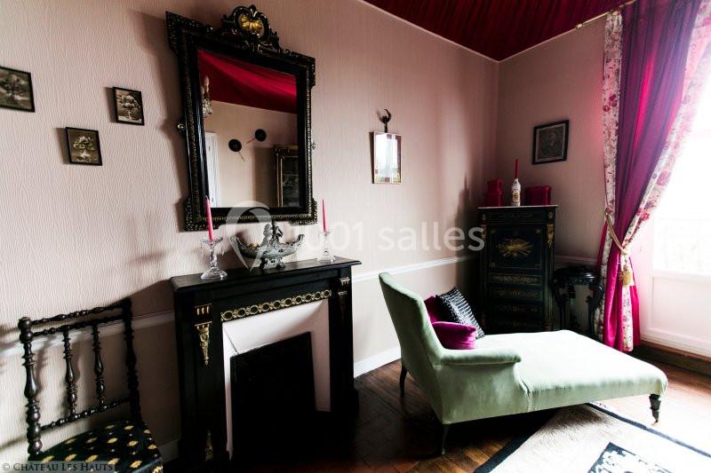 Salle élégante avec cheminée noire, miroir orné, chaise longue verte et rideaux rouges laissant passer la lumière.
