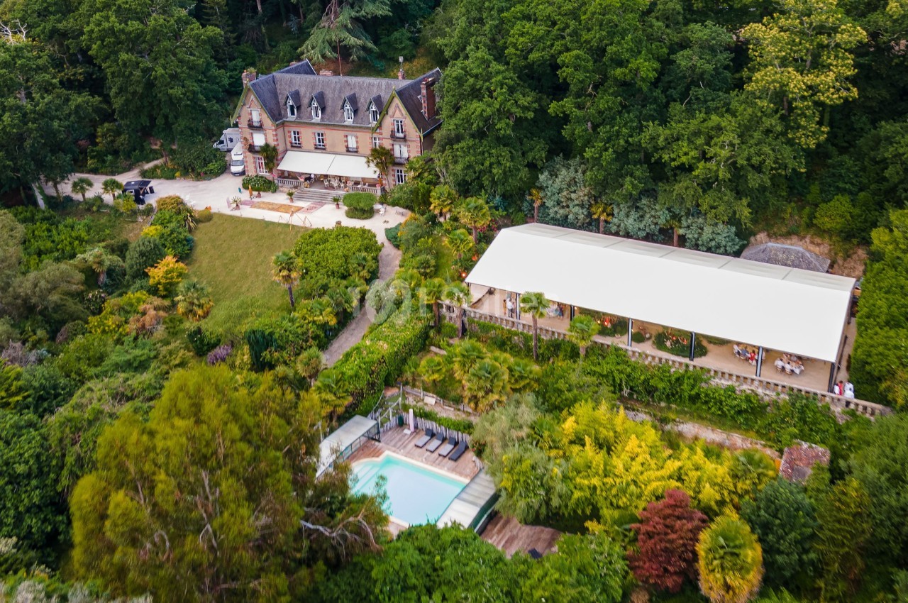 Vue aérienne d'une grande maison entourée de jardins verdoyants, avec une piscine et une terrasse couverte.