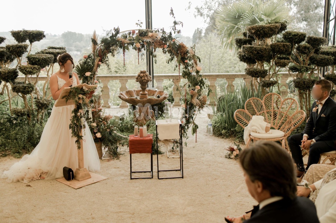 Une femme en robe de mariée lit un texte devant un autel décoré de fleurs, entourée d'invités assis.