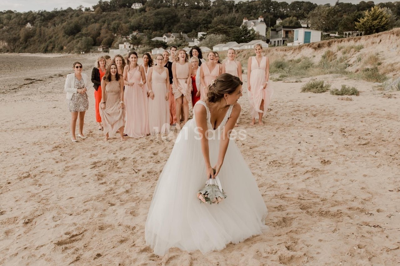 Une mariée lance son bouquet à un groupe de femmes sur une plage, avec des maisons et des arbres en arrière-plan.
