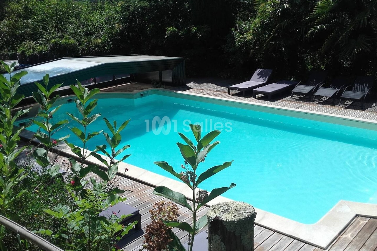 Piscine extérieure entourée de végétation, avec des transats et un abri partiellement ouvert sur le côté.
