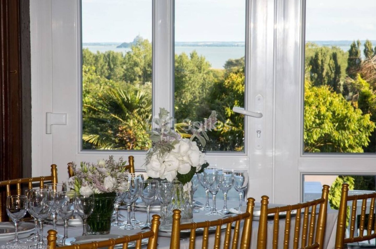 Table décorée avec des fleurs blanches et des verres, placée devant une fenêtre offrant une vue sur un paysage verdoyant et…