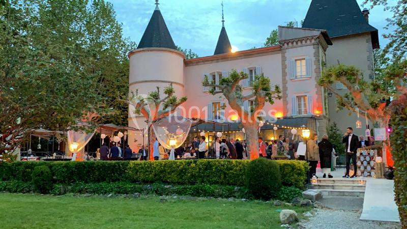 Façade d'un château éclairé au crépuscule, avec des invités rassemblés dans le jardin pour un événement.