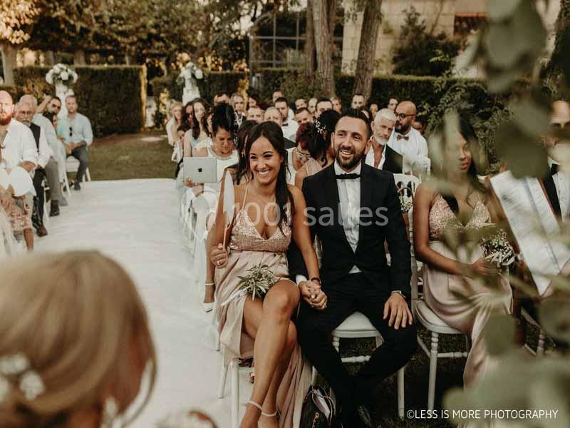 Un couple souriant assis parmi des invités lors d'une cérémonie de mariage en plein air.