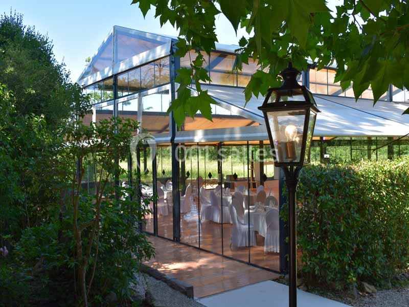 Salle de réception vitrée entourée de verdure, avec des tables dressées visibles à l'intérieur.