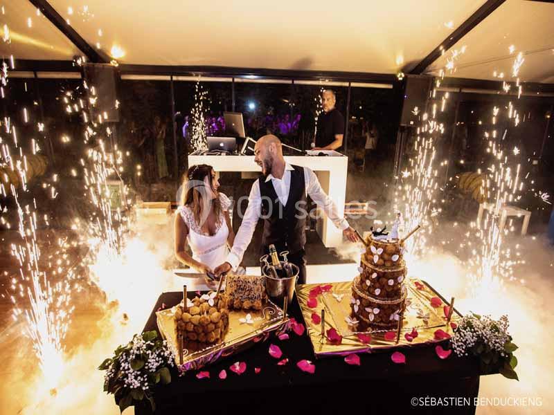 Un couple de mariés coupe un gâteau de mariage entouré de fontaines lumineuses dans une salle festive.