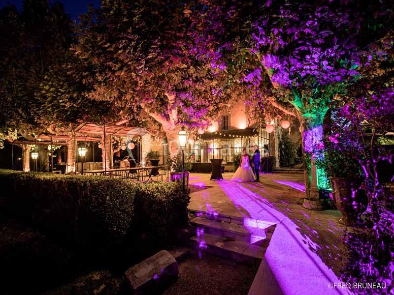 Terrasse éclairée de lumières colorées en soirée, entourée d'arbres et de végétation, avec une silhouette au loin.