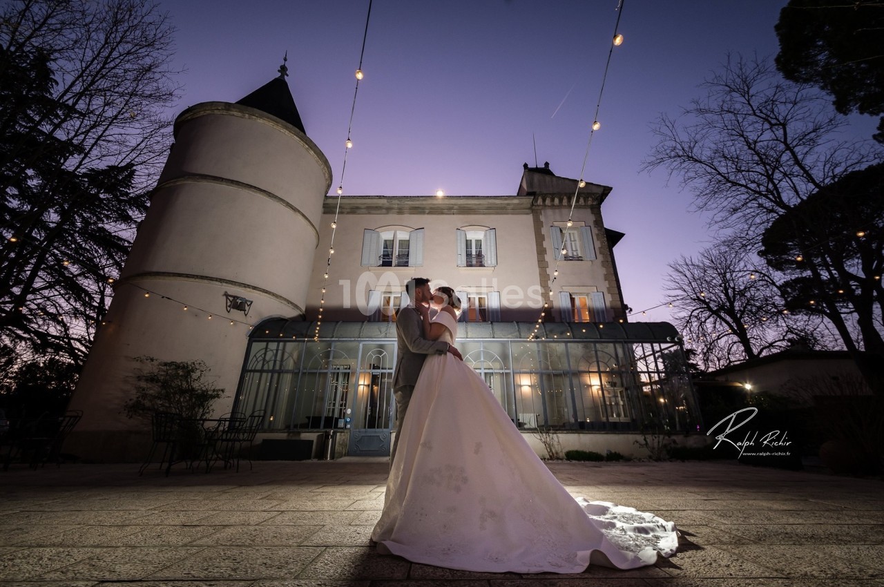 Un couple en tenue de mariage pose devant un château éclairé au crépuscule, entouré de guirlandes lumineuses.