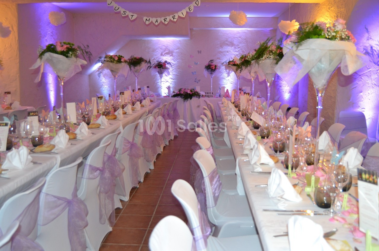 Salle de réception décorée pour un mariage, avec tables alignées, nappes blanches et ornements floraux.