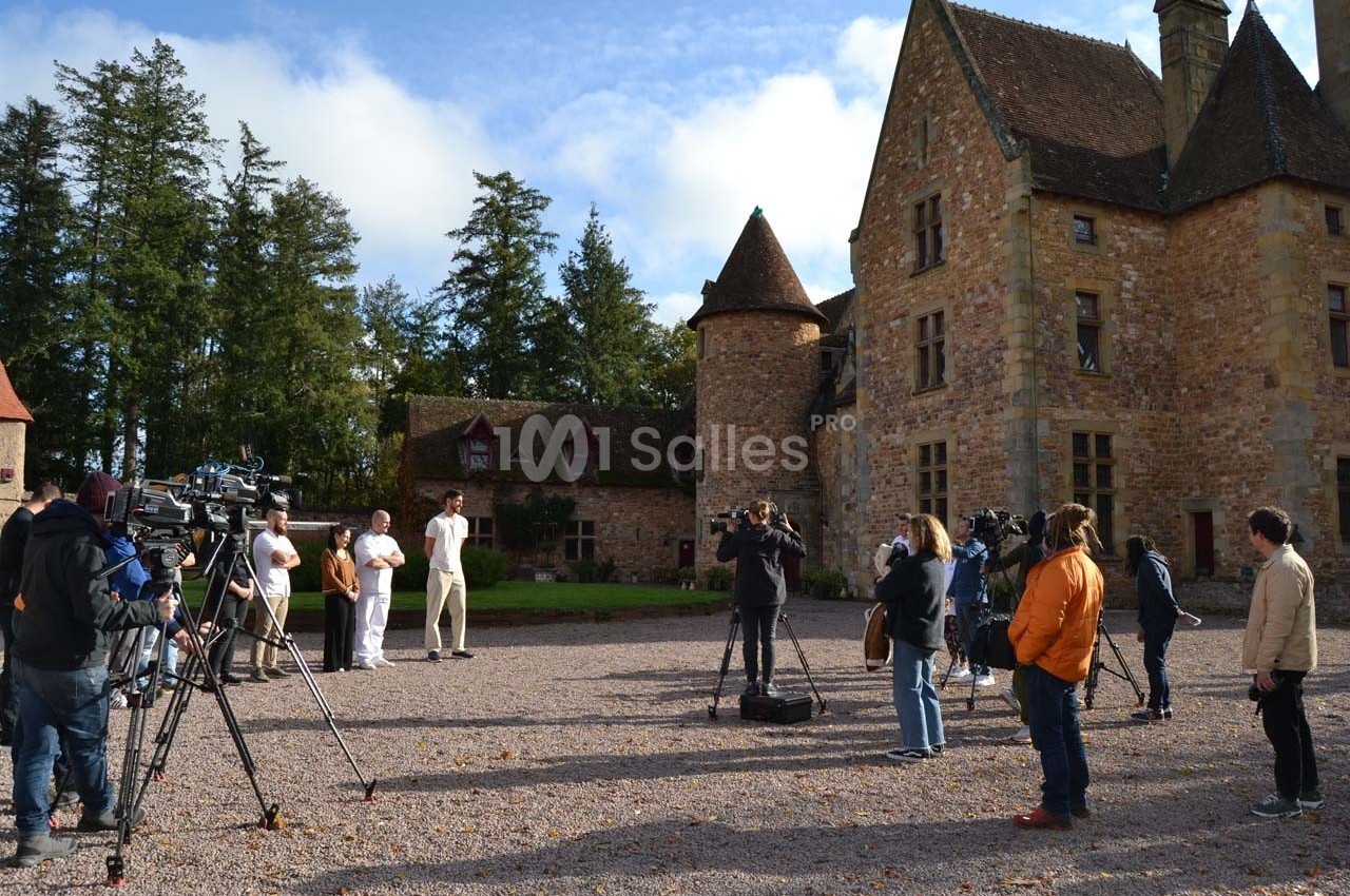 Des journalistes et caméramans filment deux personnes devant un château en pierre sous un ciel partiellement nuageux.
