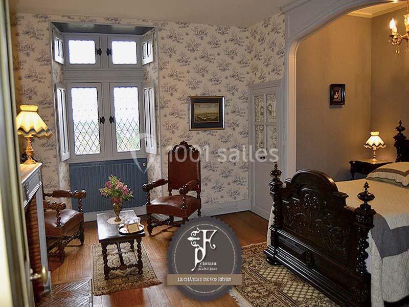 Chambre ancienne avec mobilier en bois sculpté, cheminée, fauteuils et fenêtre à vitraux donnant sur l'extérieur.