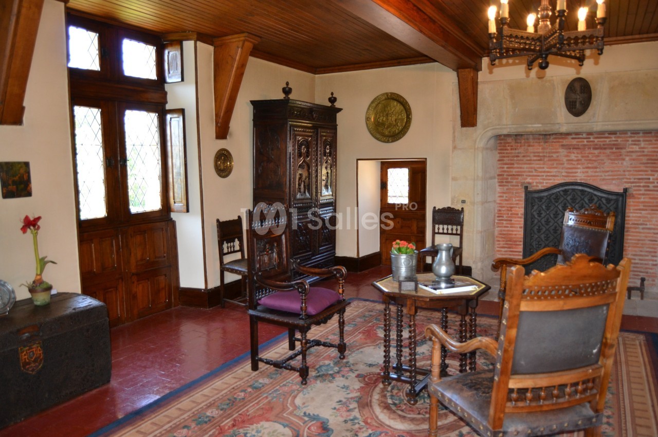 Salle de séjour ancienne avec mobilier en bois sculpté, cheminée en pierre, tapis coloré et fenêtres à vitraux.