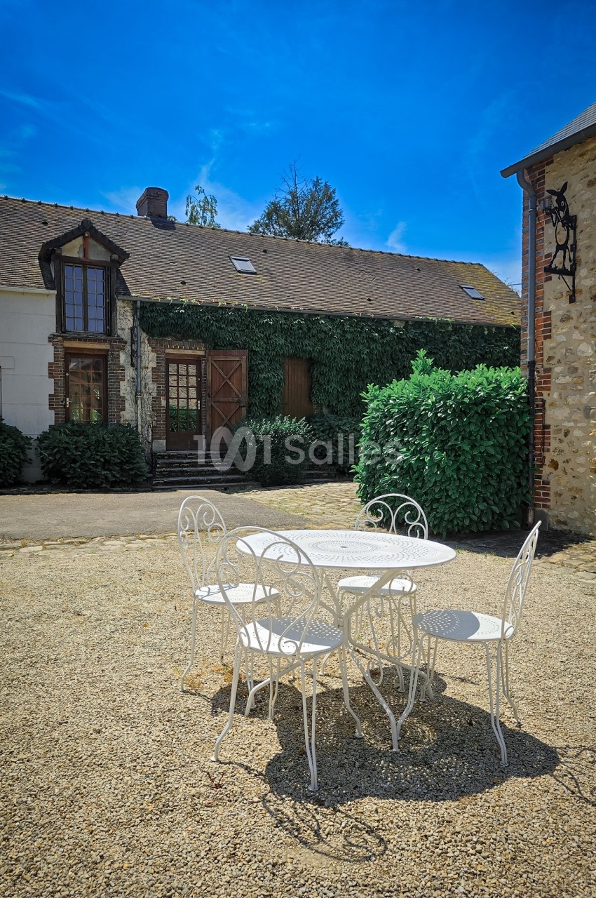 Table et chaises blanches en fer forgé sur une cour en gravier, devant des bâtiments en pierre et végétation.