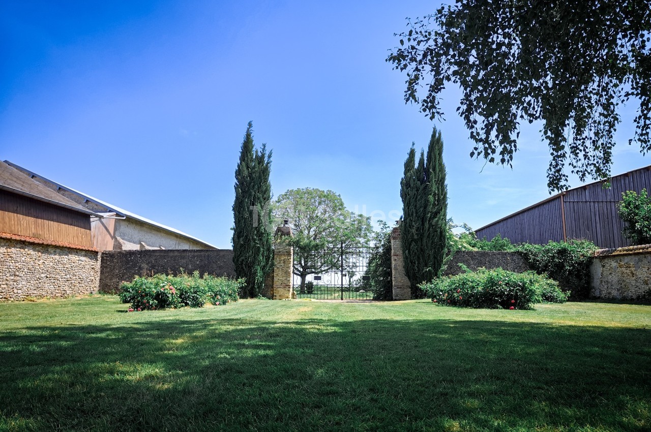 Jardin verdoyant avec pelouse, parterres fleuris, cyprès et portail en fer forgé, encadré par des bâtiments en pierre.