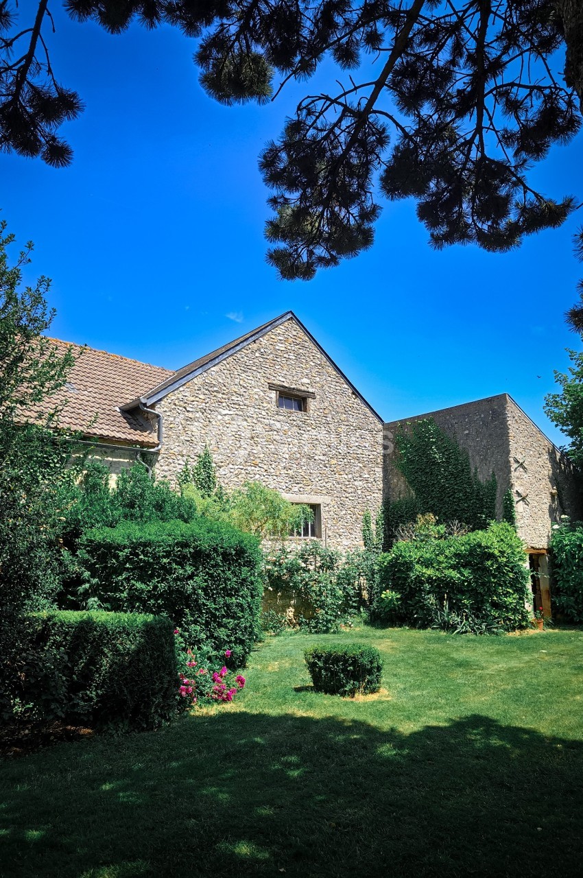 Maison en pierre entourée de verdure avec un jardin arboré sous un ciel bleu clair.