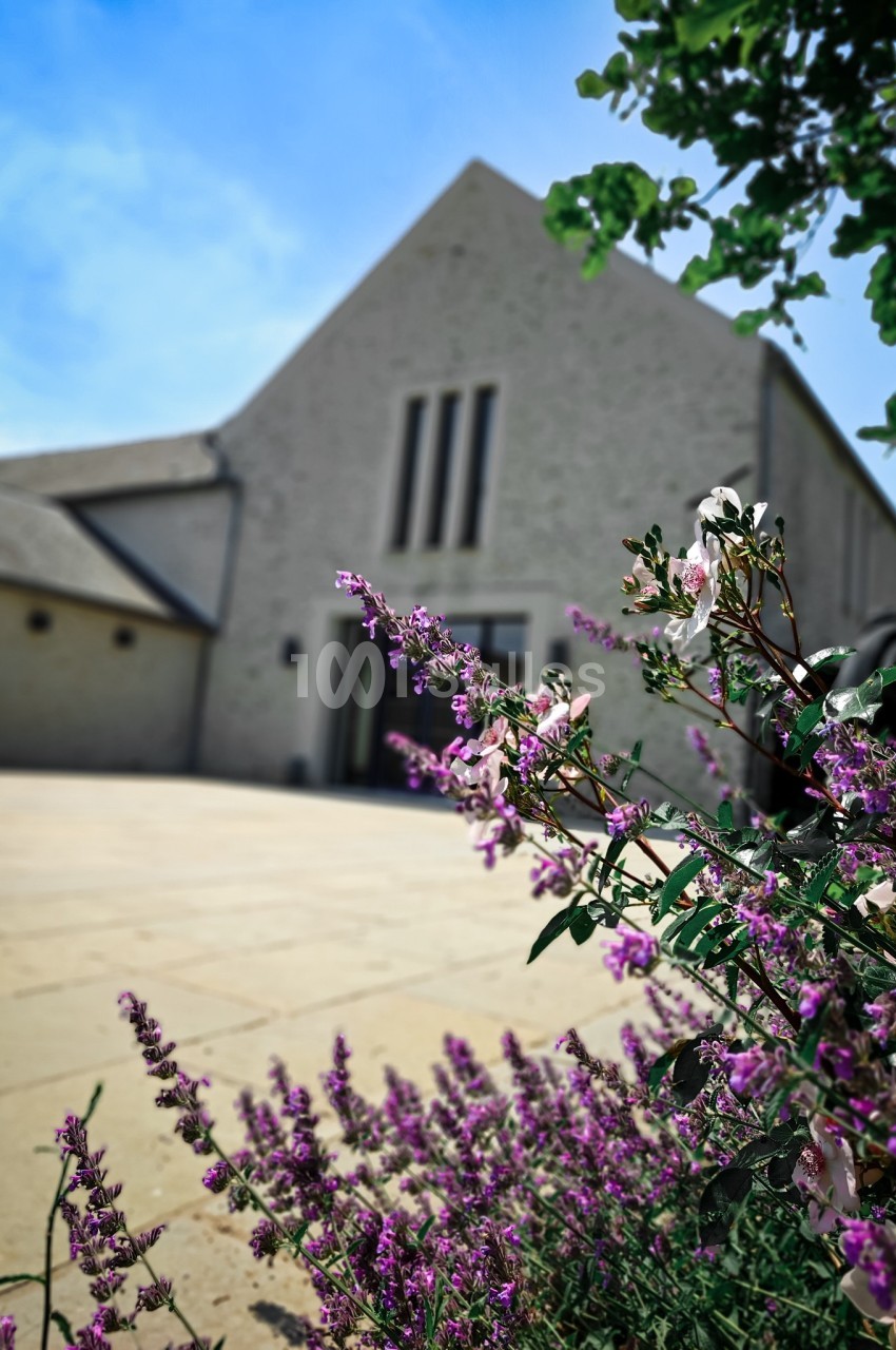 Façade d'un bâtiment moderne en pierre, entourée de fleurs violettes et blanches au premier plan.