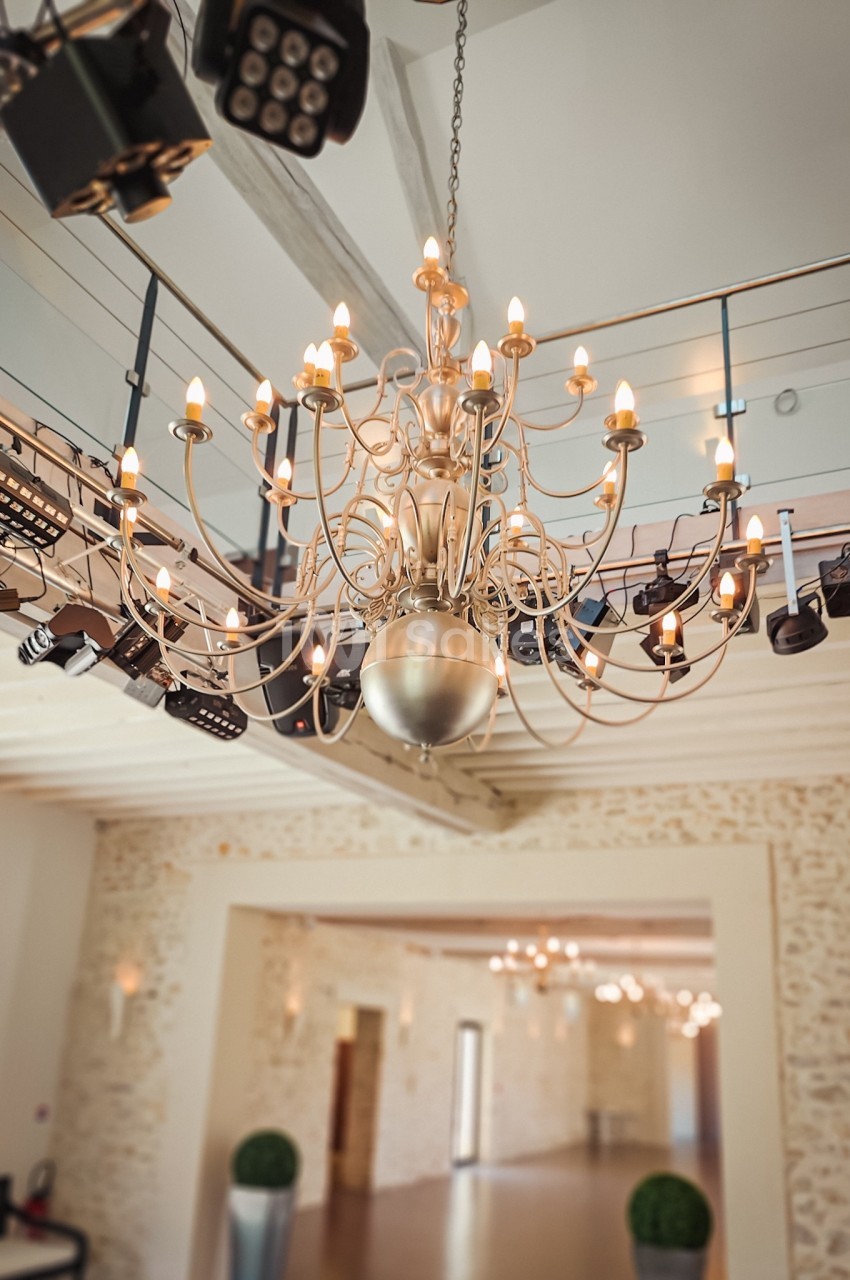 Lustre en métal avec ampoules allumées suspendu dans une grande salle lumineuse aux murs en pierre apparente.