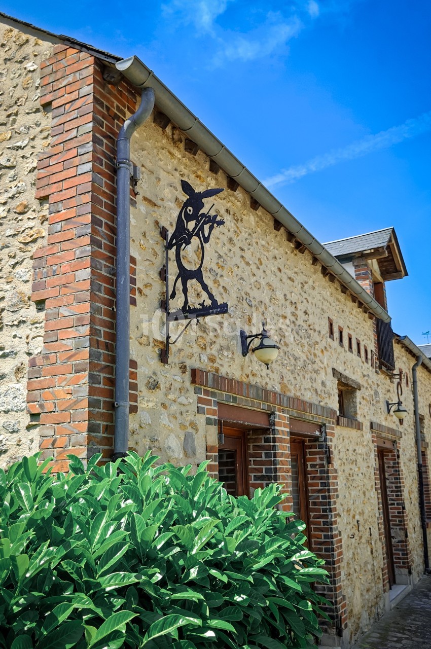 Façade en pierre d'une maison avec enseigne métallique décorative et buisson au premier plan, sous un ciel bleu.