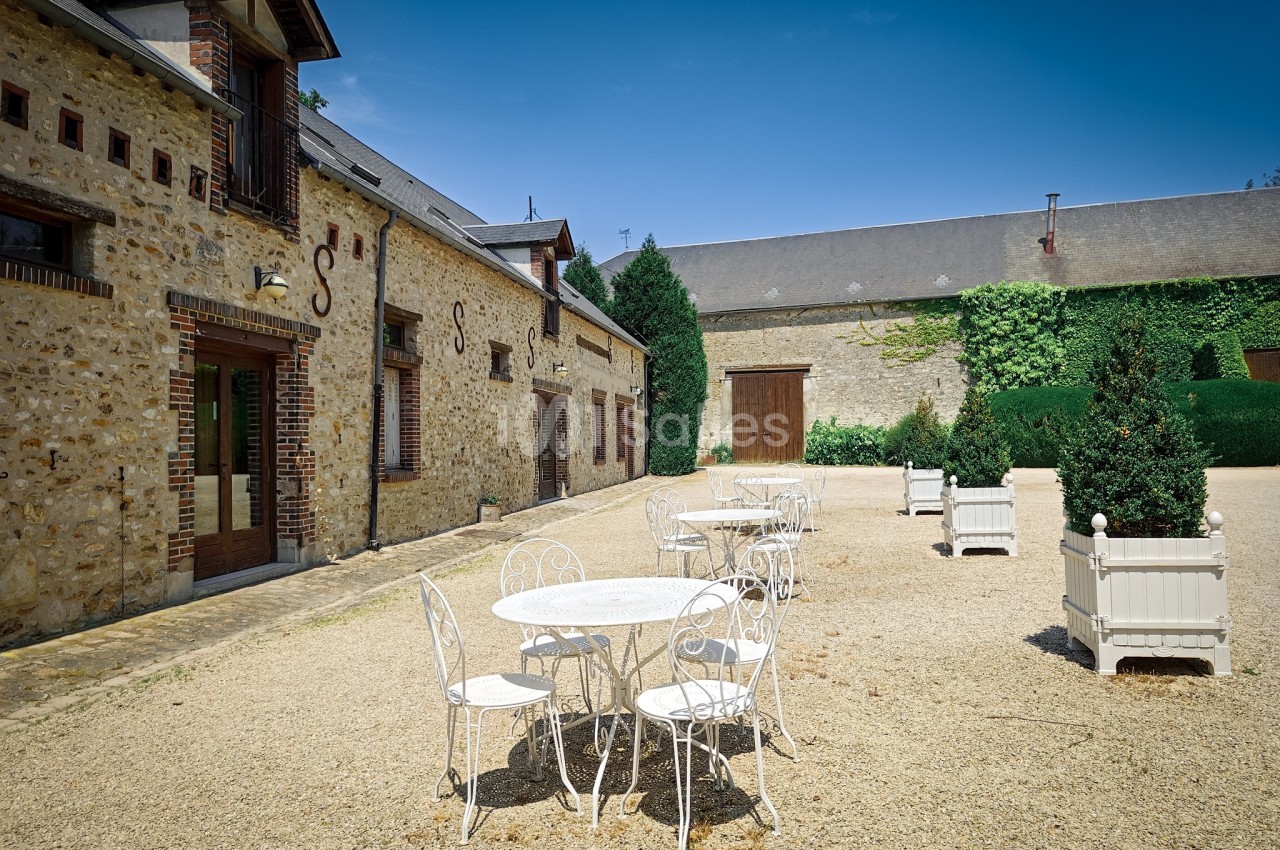 Cour extérieure avec tables et chaises blanches en fer forgé, entourée de bâtiments en pierre et de végétation.