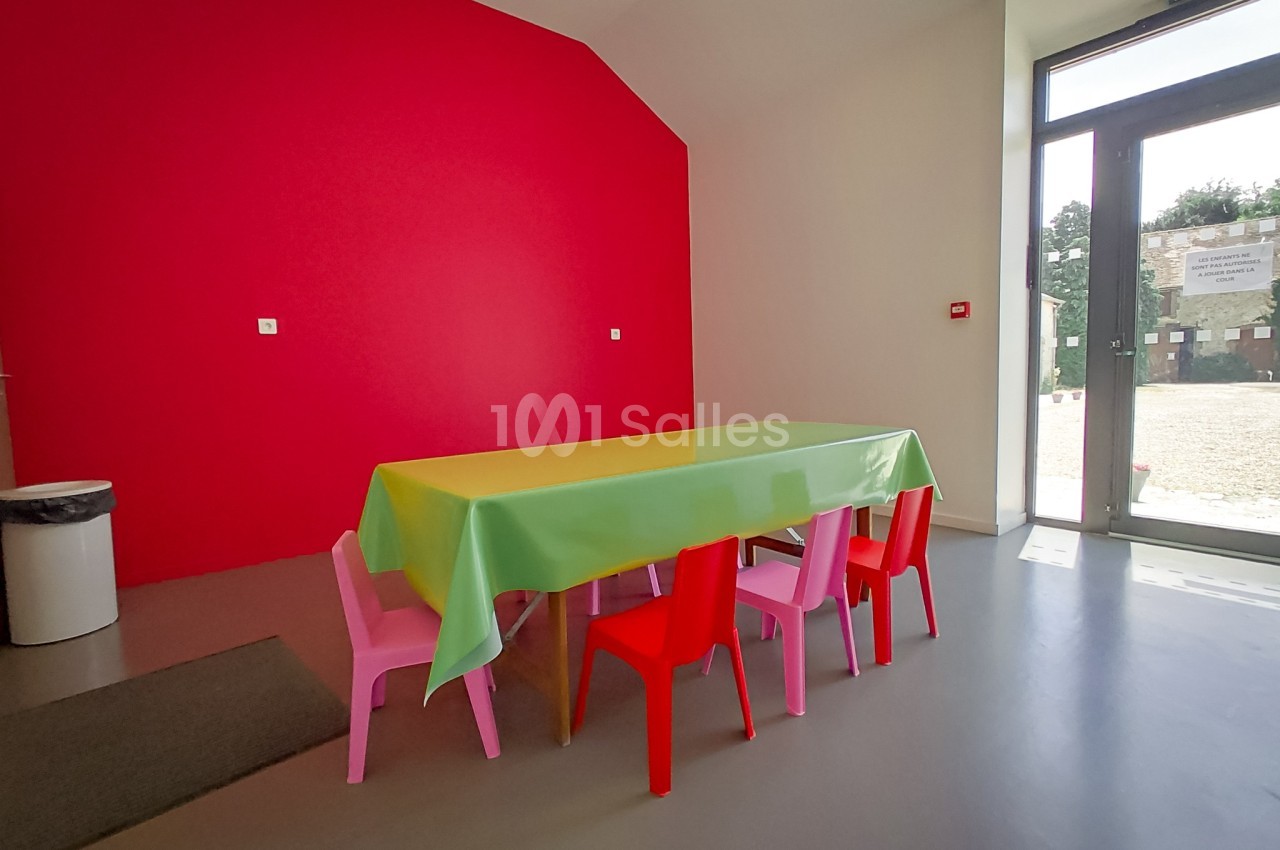 Salle lumineuse avec un mur rouge, une table recouverte d'une nappe verte et entourée de petites chaises colorées.