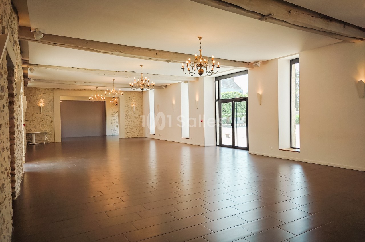 Salle spacieuse avec murs en pierre, poutres apparentes, lustres suspendus et grandes fenêtres donnant sur l'extérieur.