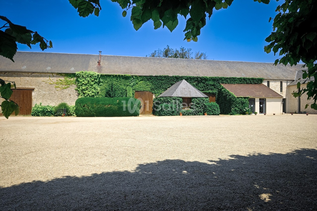 Cour en gravier devant des bâtiments en pierre avec toits en ardoise, entourés de végétation et d'arbres.
