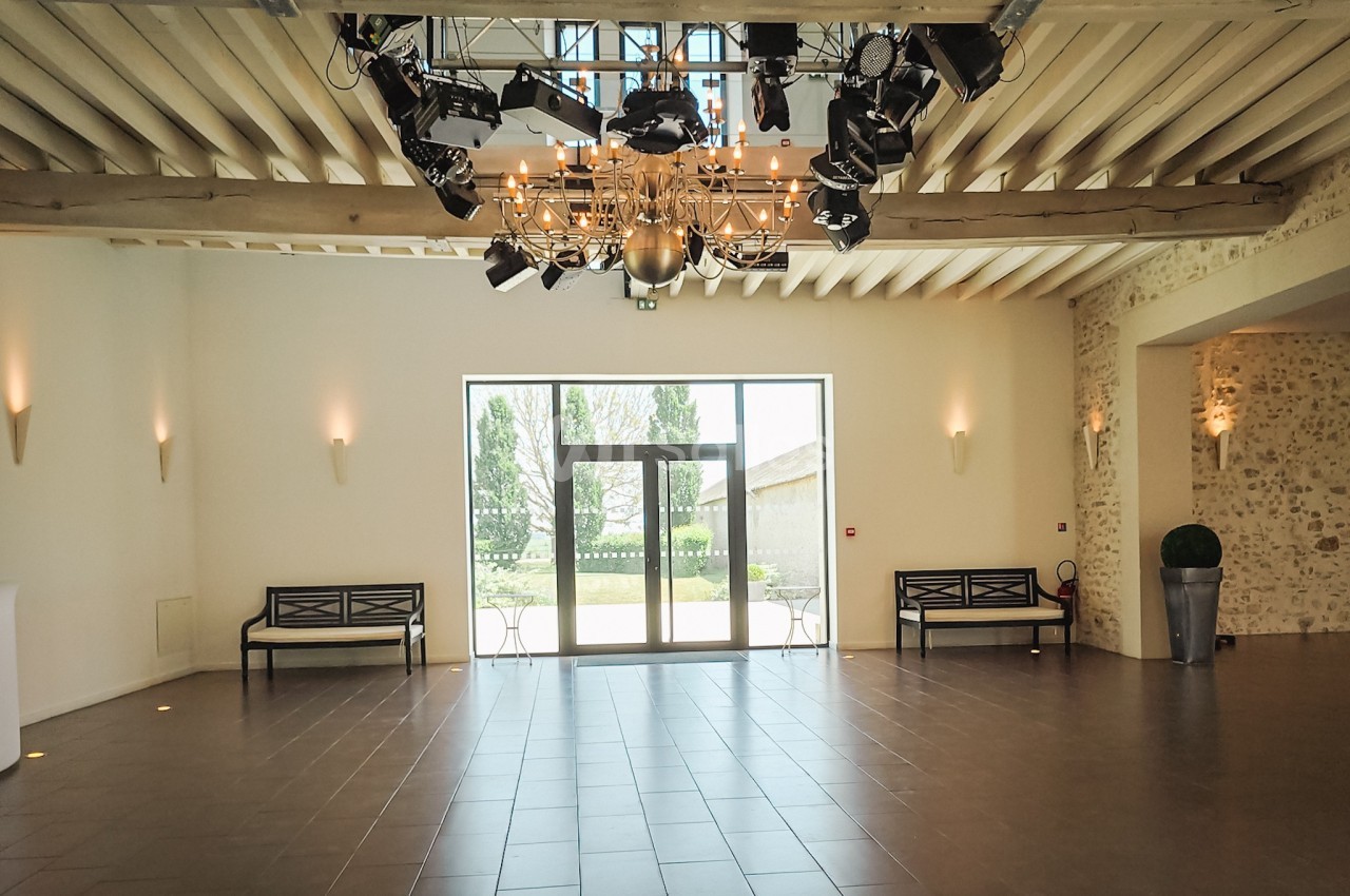 Salle lumineuse avec plafond en poutres apparentes, lustre central, bancs noirs et porte vitrée donnant sur un jardin.