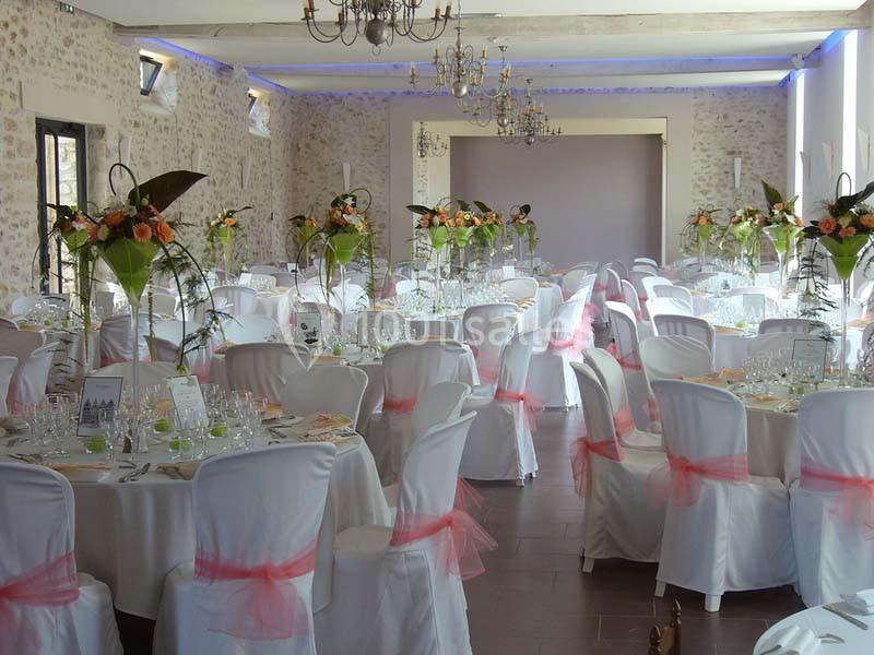 Salle de réception décorée avec des tables rondes, nappes blanches, chaises ornées de nœuds roses et centres de table…