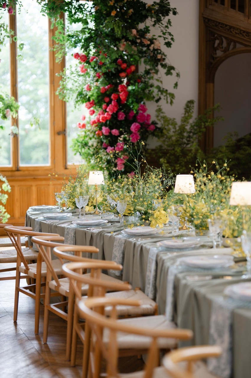 Tables élégamment dressées avec nappes vertes, chaises en bois et décor floral abondant dans une salle lumineuse.
