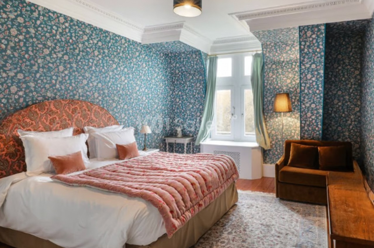 Chambre avec lit double, tête de lit rouge, murs à motifs floraux, canapé marron et grande fenêtre avec rideaux verts.