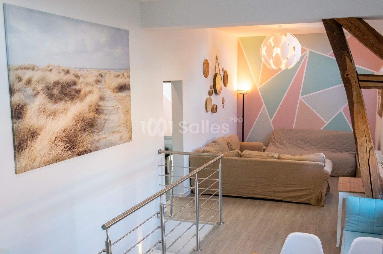Salon lumineux avec canapé beige, décoration murale géométrique pastel et tableau représentant un paysage côtier.