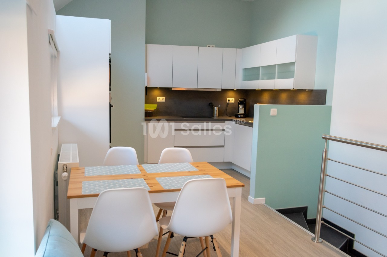 Cuisine moderne avec meubles blancs, table en bois clair et chaises blanches, éclairée par une lumière naturelle.