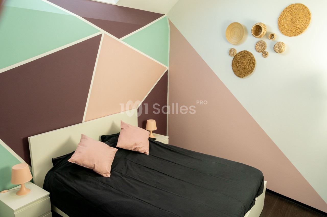 Chambre avec un mur géométrique coloré, lit double noir, coussins roses et décoration murale en osier.