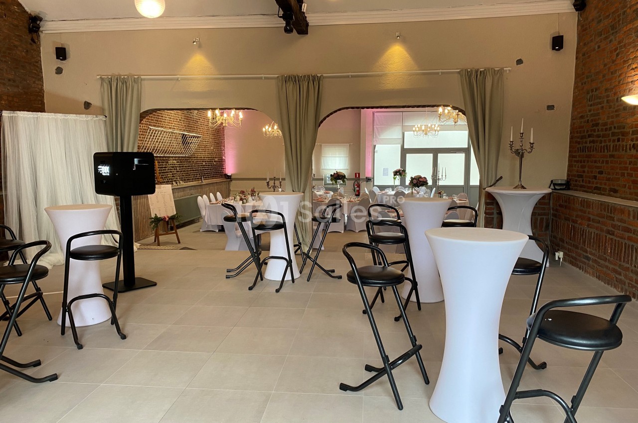 Salle de réception lumineuse avec murs en briques, tables hautes blanches et chaises noires, décorée pour un événement.