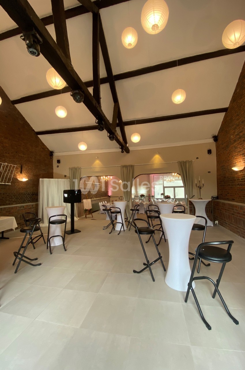 Salle de réception avec murs en briques, poutres apparentes, tables hautes et chaises pliantes sous des lampes suspendues.