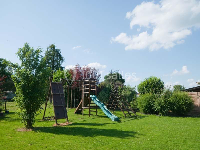 Structure de jeu en bois avec toboggan et échelle, située dans un jardin verdoyant sous un ciel dégagé.