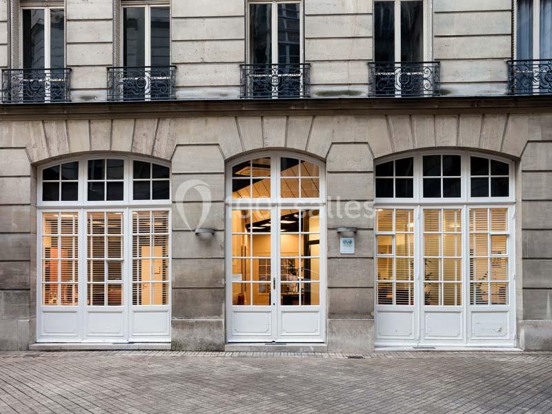 Façade d'un bâtiment en pierre avec trois grandes portes vitrées et des volets blancs, donnant sur une rue pavée.