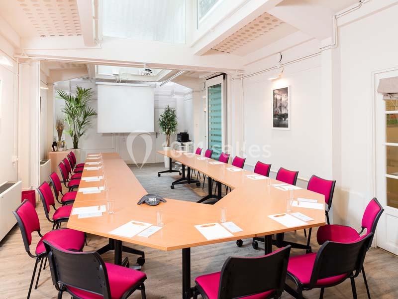 Salle de réunion lumineuse avec une grande table en U, des chaises roses, un projecteur et des plantes décoratives.