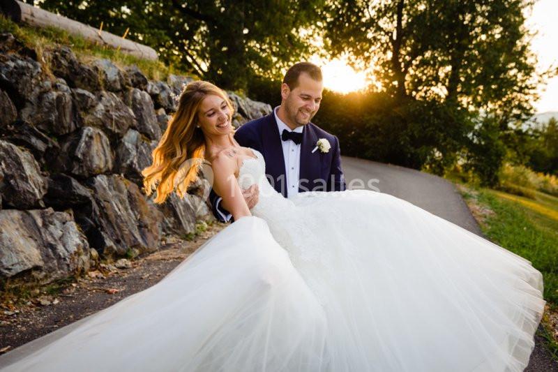 Un homme en costume porte une femme en robe de mariée sur un chemin bordé d'arbres au coucher du soleil.