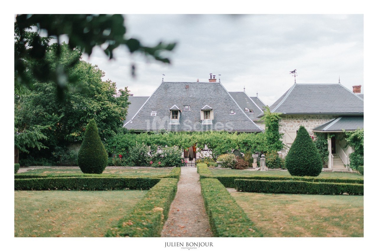 Jardin symétrique avec allée centrale menant à une maison en pierre couverte de lierre, sous un ciel nuageux.
