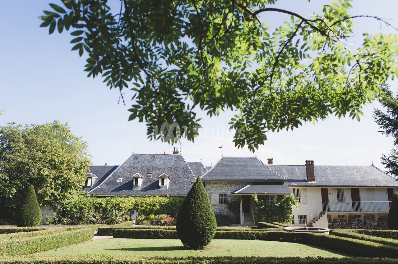 Jardin avec haies taillées et arbres, devant une maison traditionnelle aux toits en ardoise sous un ciel dégagé.