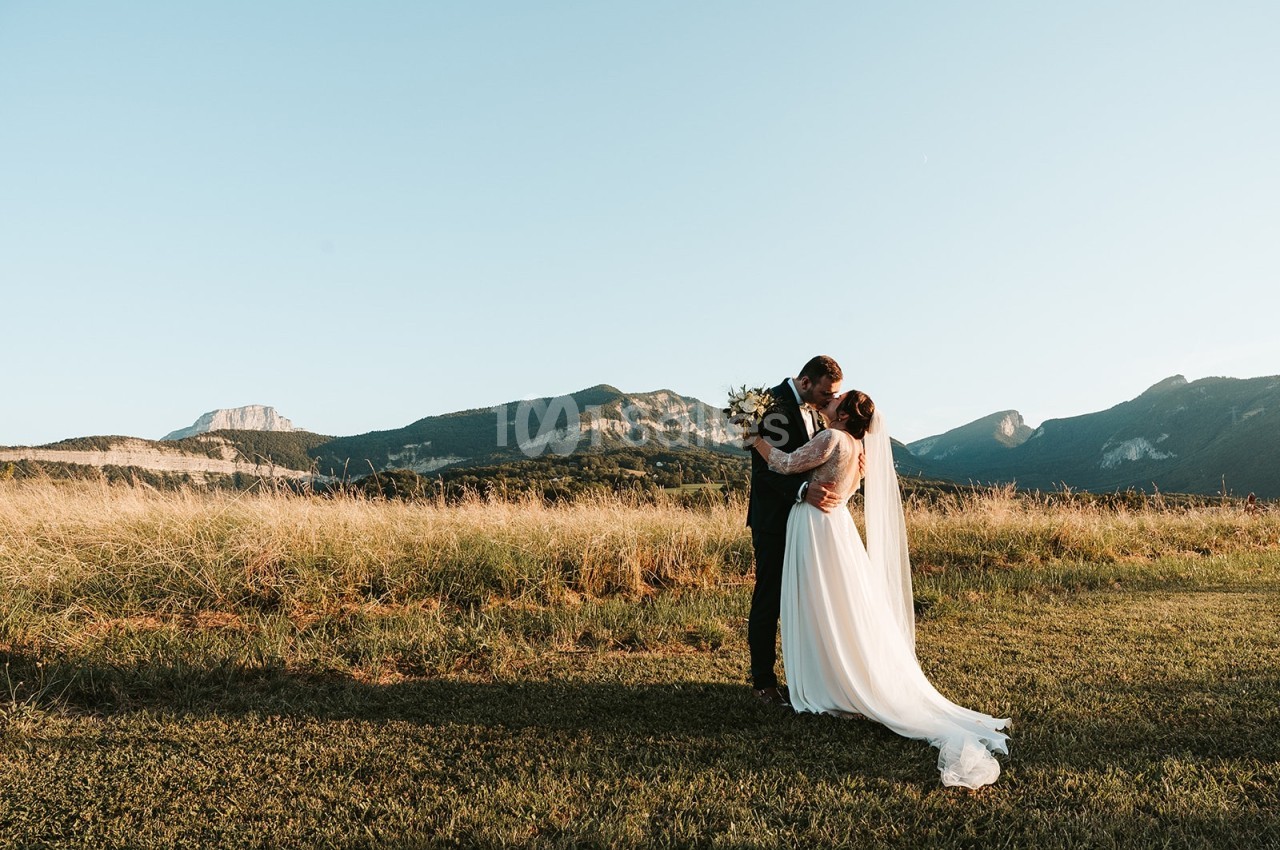 Un couple en tenue de mariage s'embrasse dans un champ au coucher du soleil, avec des montagnes en arrière-plan.